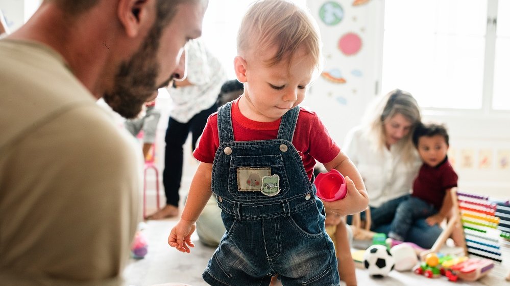 Eltern spielen in Raum mit ihren Kleinkindern, im Vordergrund ein Mann mit Kind