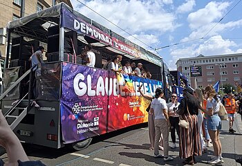Mit bunten Bannern behangener Truck mit vielen fröhlichen Menschen darauf und daneben.