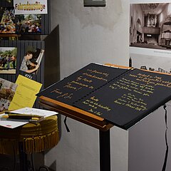 Aufgeschlagenes Gästebuch, dahinter Pinnwand mit Impressionen
