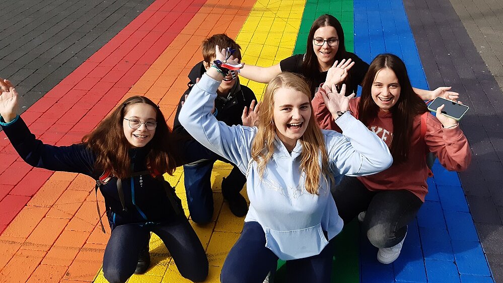 Fünf fröhliche Jugendliche Knien auf einem auf dem Boden gemalten Regenbogen