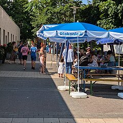 Kinder und Erwachsene sitzen auf Biergarnituren unter blauen Schirmen