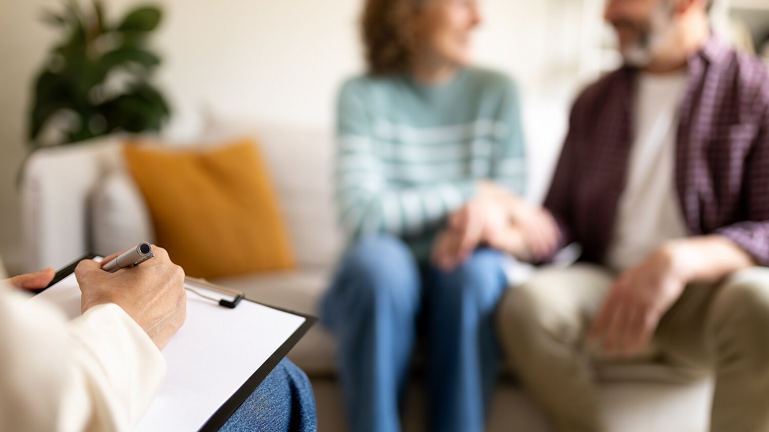 Person macht sich Notizen, während zwei Personen auf einem Sofa im Gespräch sind.