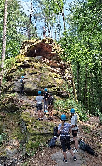 Mehrere Jugendliche mit Sicherungsausrüstung vor Felsen, von dem ein Seil runterhängt, eine Person steht auf dem Felsen
