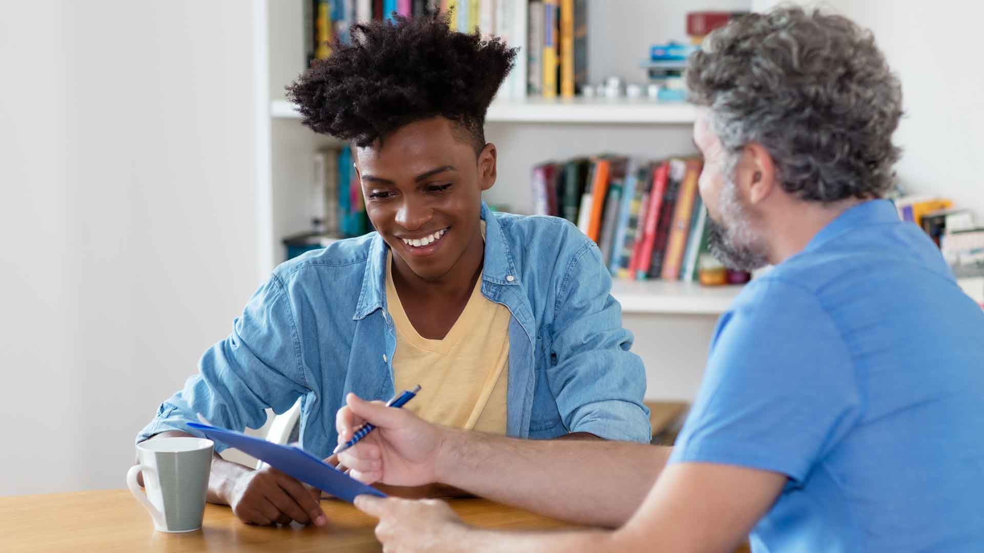 Zwei Erwachsene am Tisch; eine Person zeigt auf ein Dokument, die andere blickt darauf.