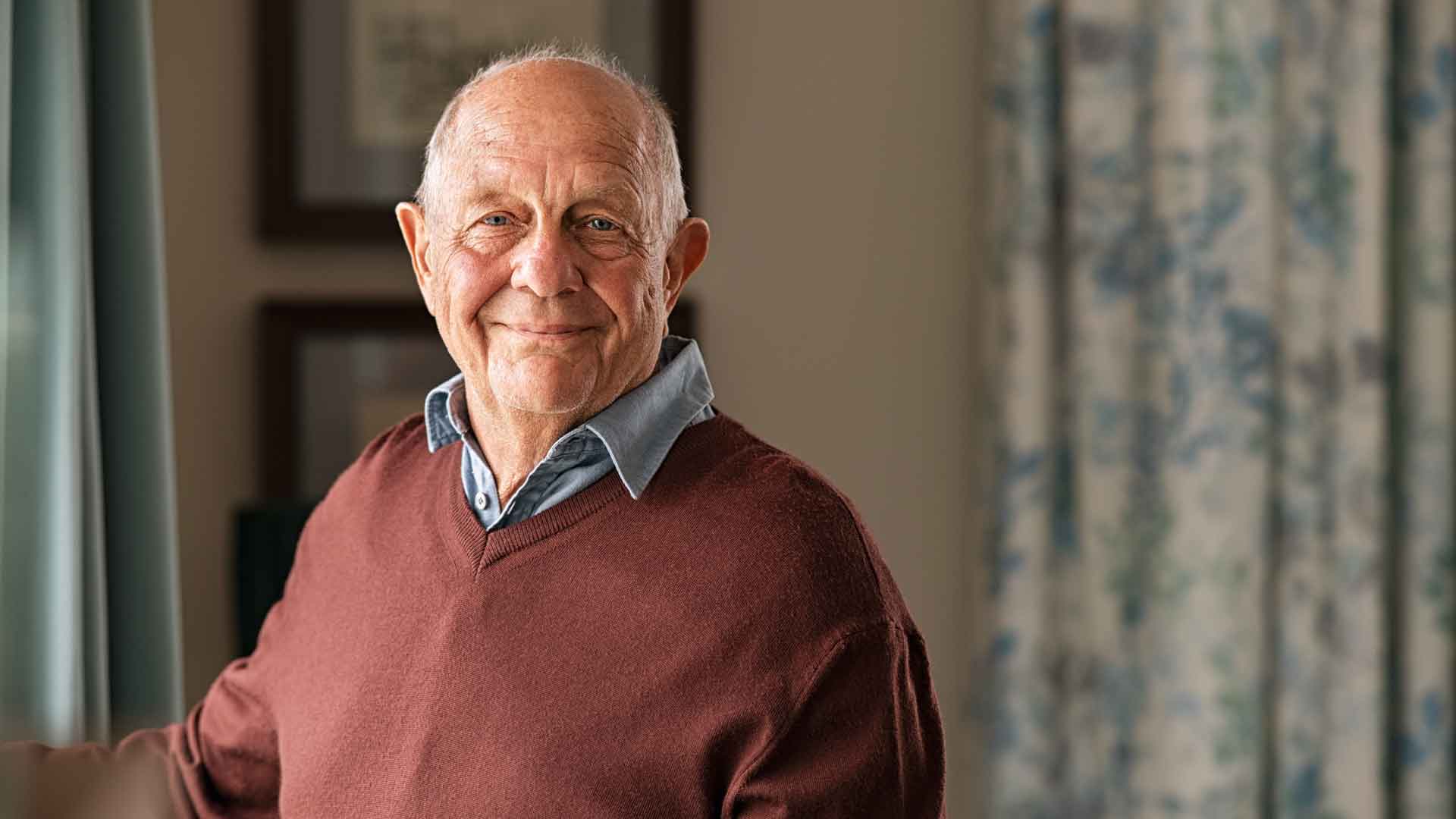 Ein älterer Mann steht in einem Wohnraum und blickt lächelnd in die Kamera.