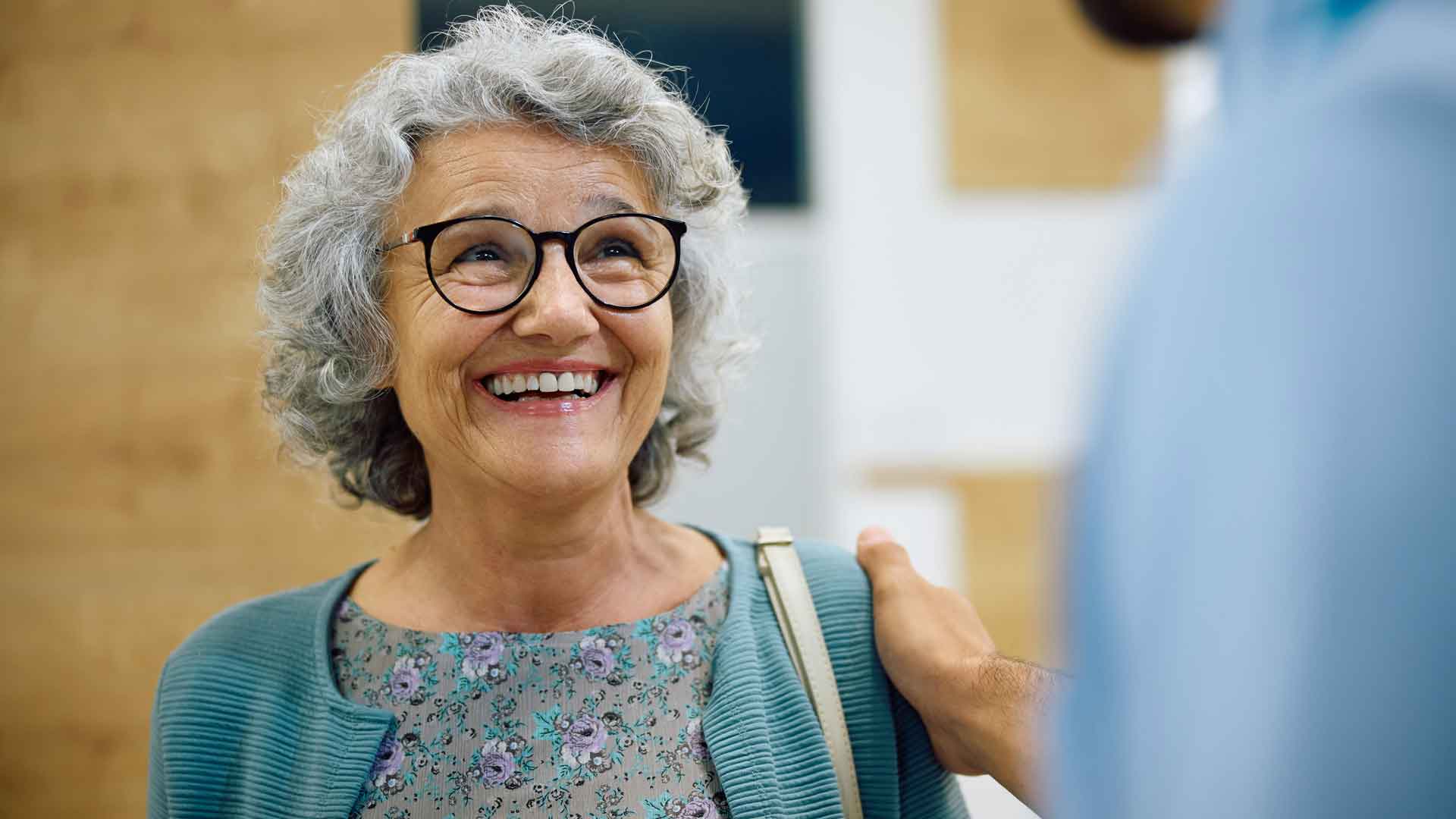 Eine ältere Frau lächelt und blickt zu einer weiteren Person, die ihr eine hand auf die Schulter legt.