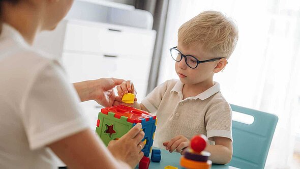 Ein Kind und eine erwachsene Person sitzen sich an einem Tisch gegenüber und spielen mit einem bunten Würfel